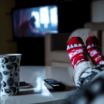 Une personne portant des chaussettes rouges à motifs se détend les pieds sur une table, à côté d'un mug, d'un smartphone et d'une télécommande, tout en regardant une télévision floue dans un salon confortable - profitant d'un moment de tranquillité avant un achat média ou naviguant sur Programmatique TV.