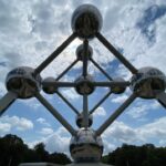 Une vue en contre-plongée de l'Atomium de Bruxelles, en Belgique, montre ses sphères métalliques brillantes et ses tubes de connexion dans un ciel partiellement nuageux. Des arbres et de la verdure sont visibles dans le bas de l'image.