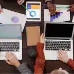 Quatre personnes travaillent autour d'une table avec des ordinateurs portables, du café et des tableaux imprimés - des mains tapent ou pointent sur des graphiques et une tablette