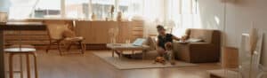 Une femme et un enfant sont assis ensemble sur le sol d'un salon moderne et lumineux, avec des meubles en bois, un canapé beige et la lumière du soleil qui traverse de grandes fenêtres. Des jouets, un ordinateur portable et l'application Wemoms sont posés sur la table basse.