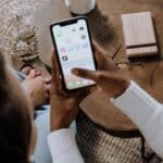 Une personne tient un iPhone et fait défiler des applications de médias sociaux, peut-être pour animer une communauté, tout en étant assise à une table basse en bois avec une autre personne assise à proximité.