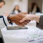 Gros plan de deux personnes se serrant la main sur un bureau contenant des documents, des tableaux et une tablette, suggérant un accord commercial entre des agences de marketing B2B Lyon. Des collègues flous sont visibles à l'arrière-plan.