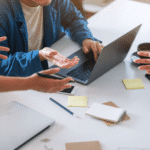 Trois personnes sont assises autour d'une table avec un ordinateur portable, des blocs-notes, des notes autocollantes et du café, engagées dans une discussion animée, un peu comme lors d'une session de création à Agences marketing B2B Bruxelles. Seules leurs mains et une partie de leur torse sont visibles.