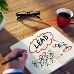 Une personne dessine dans un carnet une figure de bâton avec un mégaphone marqué "LEAD", pointant vers un groupe - illustrant comment le Top 10 des agences de génération de leads stimule la sensibilisation. Une tasse à café rouge, des lunettes et une plante en pot sont posés sur la table en bois.