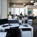 Bureau moderne à aire ouverte où plusieurs personnes travaillent à des bureaux et dans des salles de réunion vitrées, discutant de stratégies générationnelles ; de grandes fenêtres apportent de la lumière naturelle et des plantes de bureau ajoutent de la verdure à l'espace de travail.