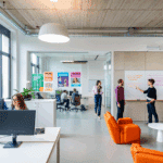 Bureau ouvert moderne avec deux personnes travaillant sur des ordinateurs et trois collègues collaborant à un tableau blanc. Les murs en verre, les affiches colorées et les chaises orange créent une atmosphère lumineuse et créative, idéale pour les agences de génération de leads à Bruxelles.
