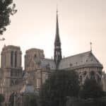 La cathédrale Notre-Dame de Paris, vue de profil, avec sa haute flèche, ses tours et son architecture gothique, à l'image des agences génération leads à Lyon qui s'élèvent au-dessus du paysage. Les arbres masquent partiellement la base, le ciel est couvert ou crépusculaire.