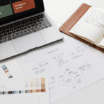 Un ordinateur portable, un carnet ouvert contenant des notes manuscrites, des échantillons de couleurs et des feuilles contenant des croquis et des diagrammes sont disposés sur un bureau blanc, reflétant un processus créatif de planification ou de conception axé sur le développement d'une ligne éditoriale forte.