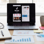 Un ordinateur portable affichant TikTok et d'autres pages web est posé sur un bureau avec une tasse à café, des écouteurs, un carnet de notes, un stylo et des graphiques imprimés - parfaits pour planifier les stratégies de marketing de Tiktok - à côté de post-it et de tableaux financiers étalés sur la surface en bois.