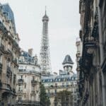 Vue de la Tour Eiffel s'élevant derrière des immeubles parisiens classiques aux façades ornées et aux balcons en fer forgé, prise par une agence de communication tourisme France par une journée nuageuse.