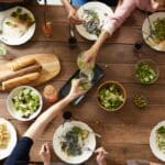 Un groupe de personnes est assis autour d'une table en bois pour partager un repas, avec des assiettes de pâtes, de la salade, du pain et des boissons. Les mains se tendent vers la nourriture et versent de l'eau, créant une scène de repas communautaire animée - un cadre idéal pour une campagne de nutrition d'une agence de marketing.