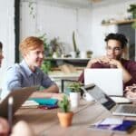 Un groupe de jeunes adultes d'une agence de marketing BtoB est assis autour d'une table avec des ordinateurs portables et des cahiers, engagés dans une discussion dans un bureau moderne et lumineux rempli de plantes.