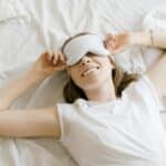 Une femme allongée sur un lit sourit en portant un masque de sommeil blanc et un t-shirt. Elle tient le masque à deux mains, profitant d'un sommeil réparateur inspiré par les conseils d'une agence de marketing santé et bien-être. Le lit est recouvert d'une literie blanche.
