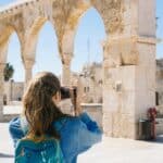 Une femme avec une queue de cheval, portant une veste en jean bleu et un sac à dos, prend une photo d'anciennes arches en pierre aux Causantes, un site historique, par une journée ensoleillée. D'autres touristes marchent à proximité.