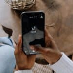 Une personne tenant un smartphone ouvert sur l'application TikTok, avec une autre personne assise à proximité. Une table en bois et un objet décoratif sont visibles en arrière-plan, ce qui suggère qu'ils sont en train de naviguer ou de faire des achats ensemble sur TikTok Shop.