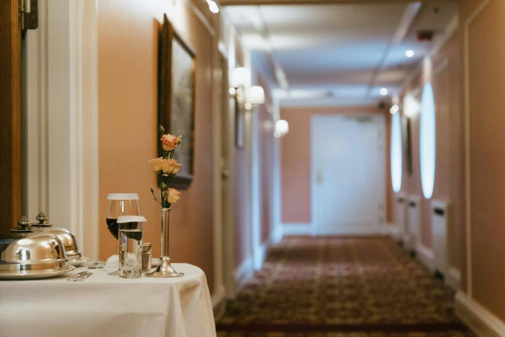 Un couloir d'hôtel avec un éclairage chaleureux et des œuvres d'art encadrées. Au premier plan, une table accueille le dîner du service d'étage - des détails qu'une agence de communication spécialiste hôtellerie en France pourrait mettre en avant pour souligner l'hospitalité exceptionnelle.
