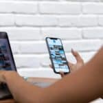 Une personne est assise à un bureau, utilisant un ordinateur portable et tenant un smartphone, parcourant les galeries de photos du contenu UGC sur les deux appareils. Un mur de briques blanches se trouve à l'arrière-plan.
